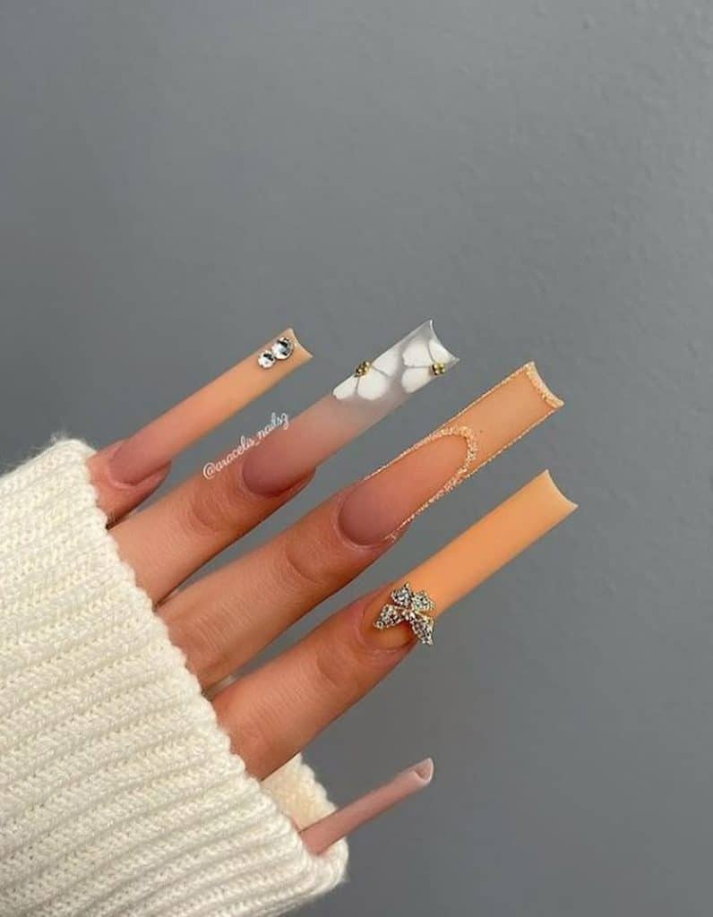 Un primer plano de la mano de una mujer con uñas acrílicas largas de color melocotón que tienen bordes, pétalos blancos, tachuelas de oro y diamantes y diseños de uñas de mariposa en 3D.