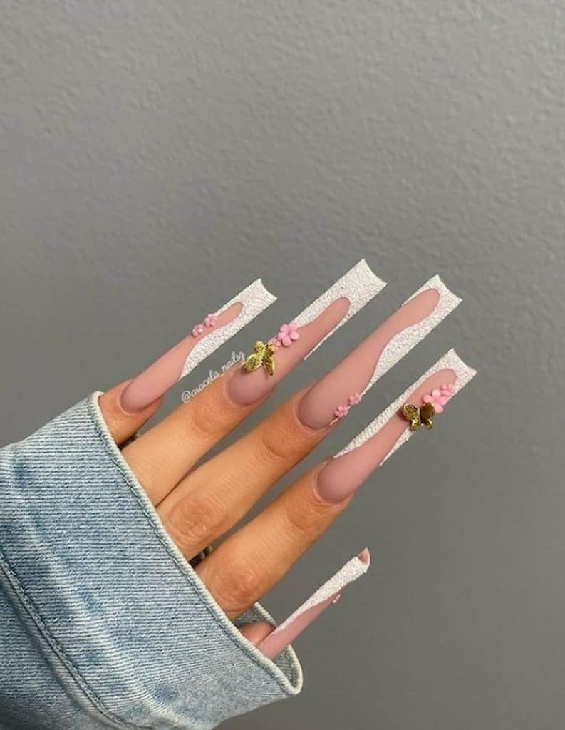 Un primer plano de la mano de una mujer con largas uñas acrílicas cuadradas y esmalte de uñas nude con brillos blancos azucarados, mariposas doradas en 3D y diseño de uñas con flores rosadas.
