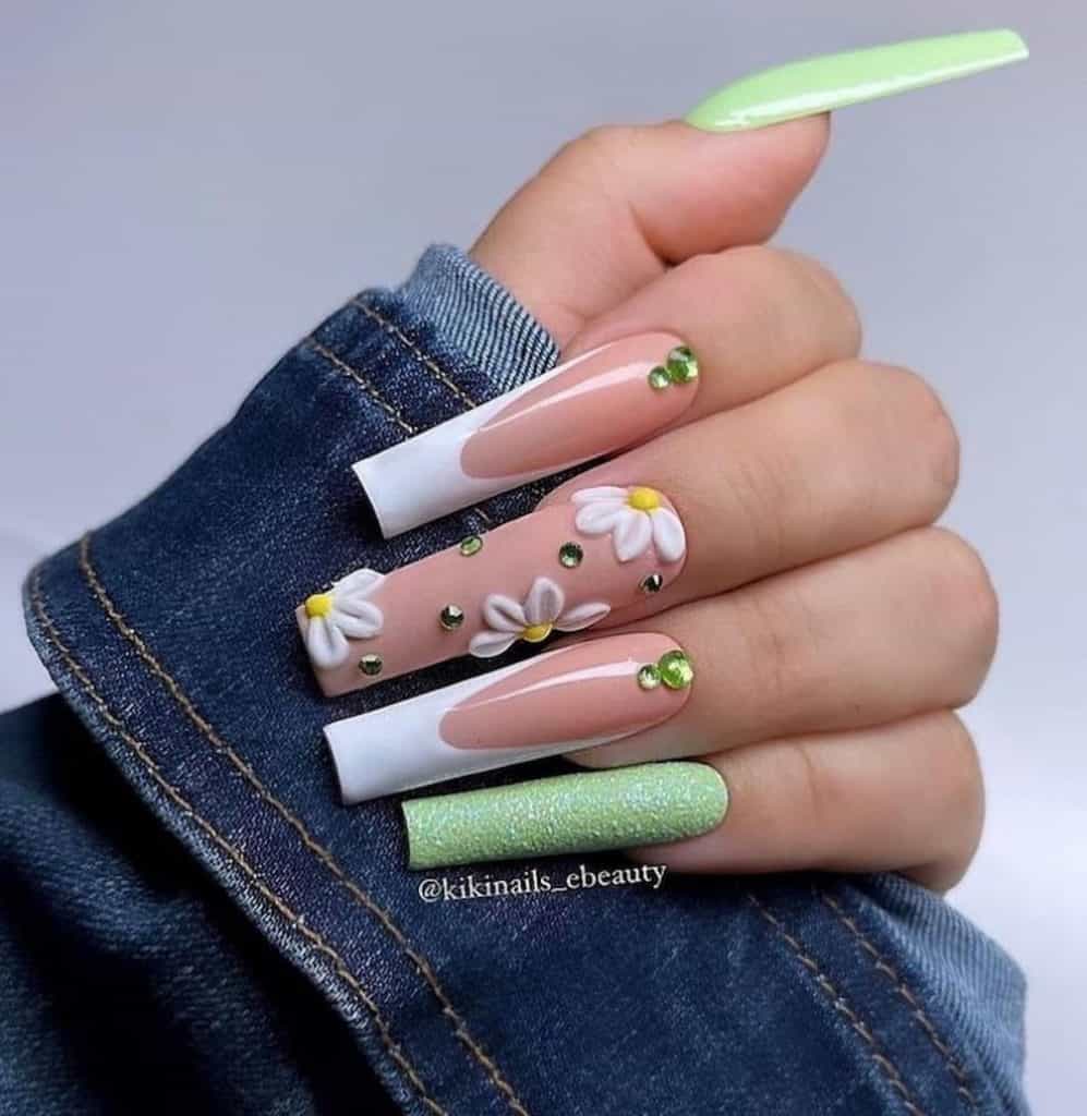 Un primer plano de la mano de una mujer con una combinación de esmalte de uñas verde y nude que tiene puntas blancas en uñas seleccionadas y diseños de uñas con margaritas en 3D y tachuelas verdes.
