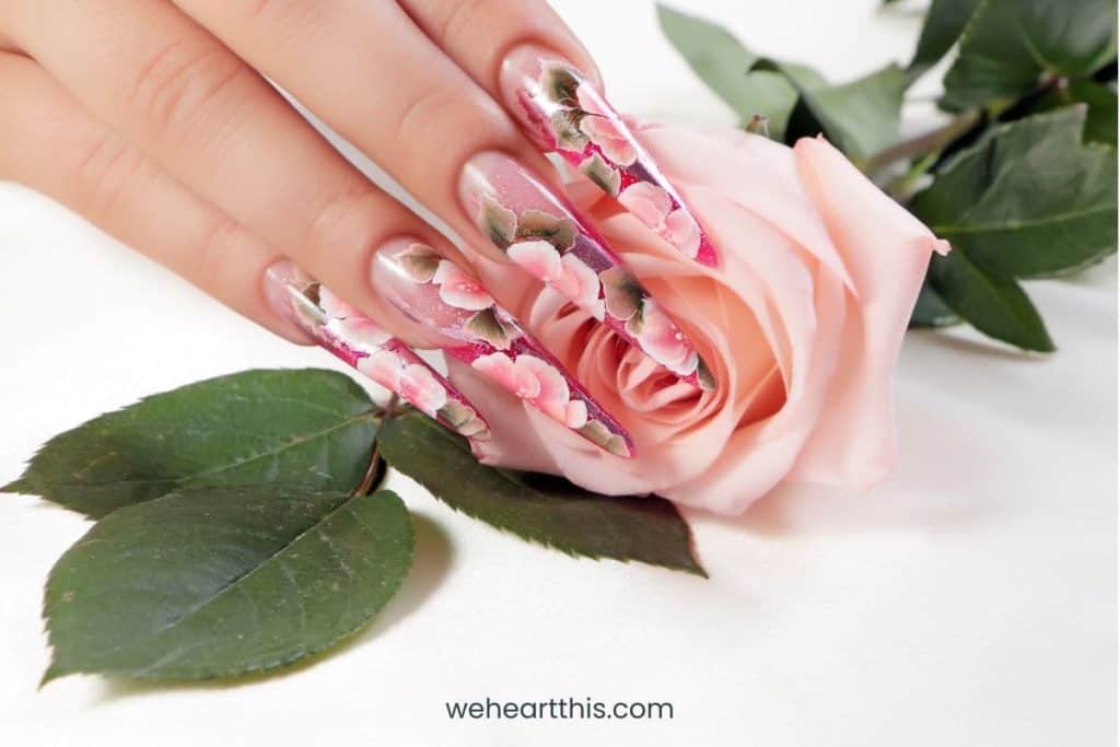 La mano de una mujer con uñas largas que tiene diseños de uñas florales, sosteniendo una rosa rosa con hojas en el fondo