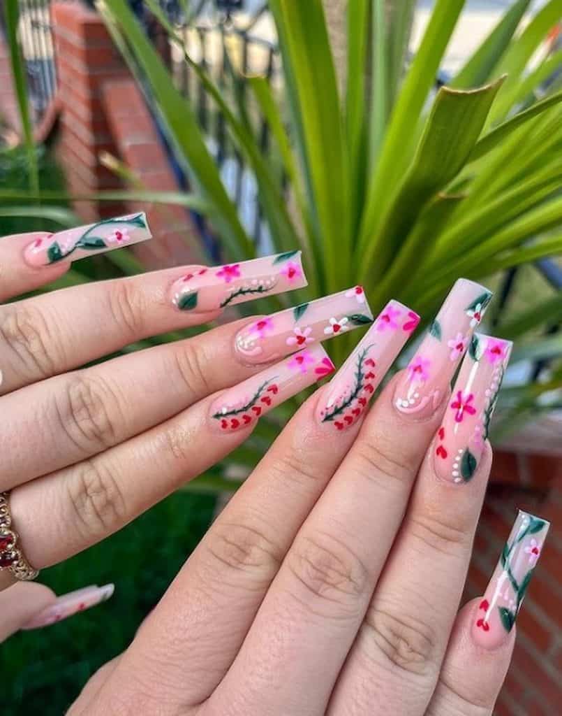 Un primer plano de las manos de una mujer con una base de esmalte de uñas rosa transparente que tiene diseños de flores, enredaderas, hojas, corazones y puntos.